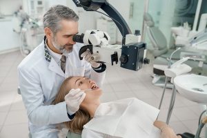 Professional stomatologist examining teeth of woman with help of modern equipment. Doctor in white coat looking through dental microscope and holding restoration instrument.